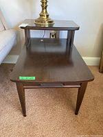 Full view of wooden side table with two tiers, lower tier larger and upper smaller with mirrored glass top piece and a brass lamp on top (lamp not included).