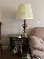 Photo showing one brass lamp with cream pleated shade on a small wooden table, next to a sofa, with a handcrafted wooden duck and goslings figurine on the table shelf below.