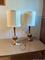 Two Mid-Century Modern style table lamps on a wood table with white fabric shades and brass and wood bases. Power cords visible and bulbs installed.