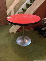 Full view of the round table showing red MDF top with black edge and metal tulip base under a staircase.