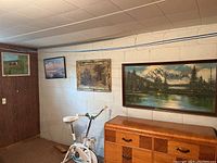 Four framed wall hangings displayed on a basement wall including horse art and landscape prints, above a wooden dresser and exercise bike.
