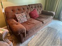 Brown tufted sofa bed with large rolled armrests, two cushions (floral and solid red), and a bright room background.