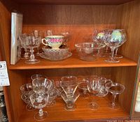 Two wooden shelves with mixed stemware items including wine glasses, snifters, fruit cups, bowls, some iridescent champagne coupes, and a floral teacup set, photographed from the front.