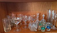 Shelf showing assortment of clear glass stemware including various stemmed glasses and small colored banded bottles with stoppers.