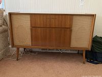 Front view showing wood veneer stereo console with left and right woven speaker covers and central drawers and fold-down door