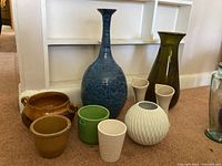 The main photo showing all nine vases and pots arranged on carpet with a white wall background, including various shapes and finishes.