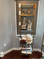 Full view of the gold metal frame shelving unit with glass shelves in a corner next to window coverings. Items on shelves include figurines and folded linens, which are not included in the sale.