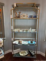 Full front view of gold metal shelf with glass shelves filled with vintage-style collectibles including porcelain dolls, cups, plates, and other decorative items.