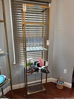 Full view of tall gold metal frame shelving unit with four glass shelves against window with blinds.