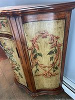 Side view showing hand-painted decorative motif on side panel with reddish vine scrolls and green leaves on a yellow background, framed by warm brown wood edges.