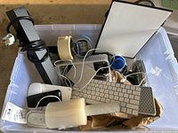 Overview of tote showing various items including Apple keyboard, Magic Mouse, power strip, packing tape and assorted cords and accessories inside clear plastic container.