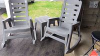 Two gray plastic rocking chairs with slatted backs and one gray plastic side table. Outdoor setting with visible driveway and grass area.