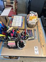 Photo of various pens, markers, plastic palette, rulers, business card sleeves, and other art supplies on a cardboard surface.