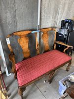 Front and side view of a solid wood antique double seater bench with red seat cushion positioned indoors against a wall.