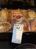 Top view showing two ornate metal cups with lids and saucers, a package of Neyba Turkish coffee, and a long decorative spoon.