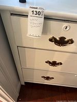 Close-up photo showing white dresser with brass handles and label with lot number and description.
