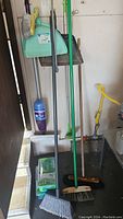 Photo showing multiple push brooms with long handles and a blue dustpan hung on wall, plus a cleaning bottle.
