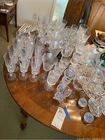 Photo showing a large variety of crystal and glass stemware on a wooden table including champagne flutes, wine glasses, and decorative items.