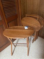 Four wooden folding dinner trays with oval tops stacked against a wall showing their size and wood finish.
