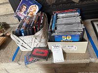 Photo showing a basket and box containing DVDs, CDs, and a cassette tape from varying genres along with visible titles and the lot label.