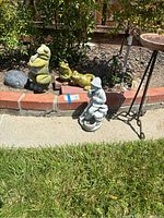 Photo showing the larger frog playing accordion, two smaller frog statues, and the man statue with plant holder all placed on a brick garden border and paved lawn edge.