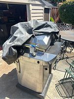 Full view of Char-Broil gas grill with black cover partially draped over it. Grill is stainless steel with cabinet base and side shelves.