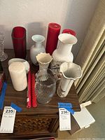 Photo showing a collection of various vases in white ceramic and clear glass, along with several red candles placed on a wooden surface.