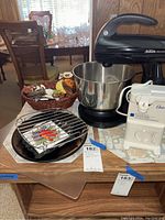 Photo of Black Sunbeam Mixmaster stand mixer, Oster white electric can opener, cast iron cookware including a drip pan and grill rack with a decorative tile, and a wicker basket of owl-themed decor items on a wooden table.