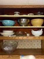 Full shelf view showing multiple kitchen bowls and plates arranged on wooden shelving with floral shelf liner.