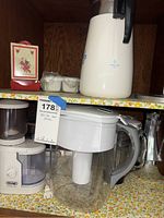 Shelf with Brita water filter pitcher, white electric food chopper, white coffee carafe or thermos, and other kitchen items.
