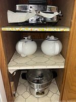 View of the Black & Decker waffle iron on top shelf with yellow floral patterned trim; two white ceramic kettles with blue accent lids on middle shelf; stainless steel automatic popcorn maker and steam pot on bottom shelf.