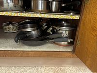 Photo showing stack of metal pots and pans with black handles inside wooden cabinet, including a casserole dish with lid featuring green floral pattern.