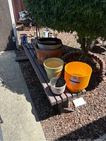 Outdoor bench with assorted plant pots arranged on and around it: large yellow plastic pot, green plastic pot, various terracotta pots, and textured ceramic pots. Metal plant hangers are also visible.
