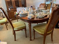 Oval wooden dining table surrounded by four matching wooden chairs with cane backs and green fabric seats. Table top covered with various dishes and glassware.