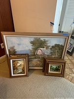 Three framed prints leaning against the wall on carpet. Large landscape with cottage and two smaller landscape prints with forest and river scenes.