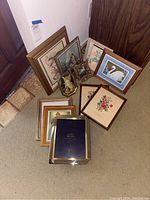 Photo showing a collection of around 10 framed prints placed on a carpet and leaning against a wall. Includes bird and floral themed prints along with a small framed figurine.