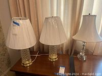 Photo of three table lamps placed on wooden table under warm lighting with cream curtains in background.