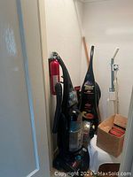 Closet corner showing two Bissell upright vacuums, fire extinguisher, mop/broom handle, white stool, and a box of lightbulbs.