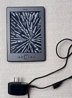 Amazon Kindle 4th generation e-reader with charging cord shown laying on a light-colored fabric surface. Screen shows a decorative image of many fountain pen nibs.