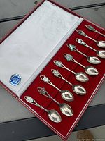 Open case showing 12 silver spoons with decorative handles, red velvet interior, and white cloth inside lid
