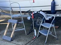 All items in the lot shown outdoors on concrete ground, including two metal foot stools, a rolling dolly, adjustable work platform, and drain snake