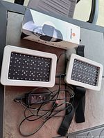 View of LED light therapy device with two rectangular panels, black LED bulb surfaces, white borders, attached straps and power adapter.