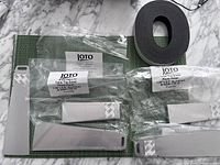 Photo showing multiple bags containing metal table top easels labeled JOTO with 3M adhesive backing, aluminum material, two sizes visible, and a large roll of black heavy duty magnet tape.