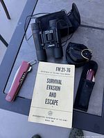 Photo showing the complete lot on a table: binoculars, compass, Swiss Army multi-tool cutter, and survival manual.