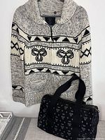Front view of TNA Aritzia women's sweater with zip front and geometric knit pattern, next to black TNA handbag with logo print