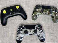 Three wireless PlayStation controllers on gray fabric surface: black controller with yellow thumbsticks, green camo controller, gray camo controller