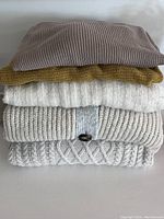 Stack of five sweaters showing various knit patterns and neutral colors.