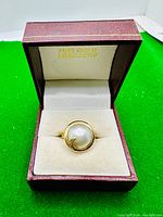 Ring displayed in open burgundy jewelry box with cream interior, showing front of ring with pearl and gold crescent surrounding it.