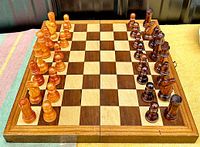 Complete wooden chess set arranged on the board with carved light and dark wood pieces in starting position. Board is wooden with square grid, stored pieces inside
