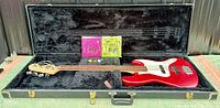 Wide shot of red Fender Squier Jazz Bass inside open black hardshell case showing entire guitar, case interior, cable, string packs and picks.